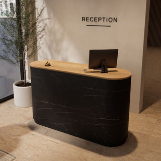 A modern curved reception desk with a black marble-look front and a light wood top, placed in a lobby setting with a computer monitor, bell, and a potted plant nearby. The wall behind has the word 'RECEPTION' mounted on it.