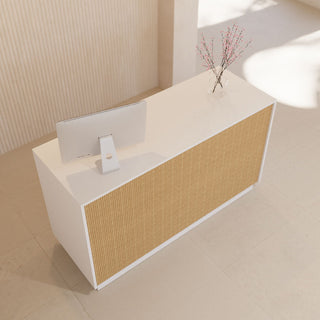 A modern white reception desk with a textured beige ribbed front panel, styled with a computer monitor and a small vase of branches, set in a minimalist beige interior.