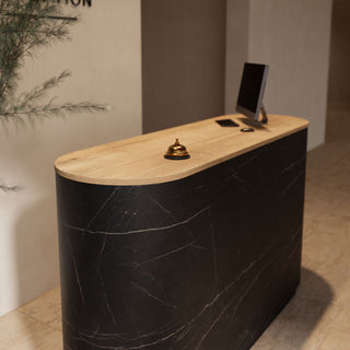 A modern curved reception desk with a black marble-patterned body and a light natural wood top, styled in a lobby setting with a computer monitor, phone, and brass bell on top.