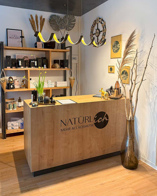 A modern wooden reception desk with the brand name 'Näturlích' printed on the front, set in a styled cosmetics shop interior with shelves, decorative dried grass, and warm lighting.