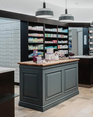 A modern pharmacy counter with a dark gray paneled base and a light wood top, set in a brightly lit retail space with shelves of medicine and hanging pendant lights.