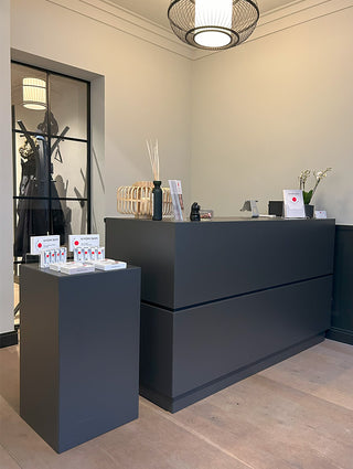 A modern minimalist reception desk and matching display pedestal in matte dark gray, set in a clean boutique interior with beige walls and stone flooring.